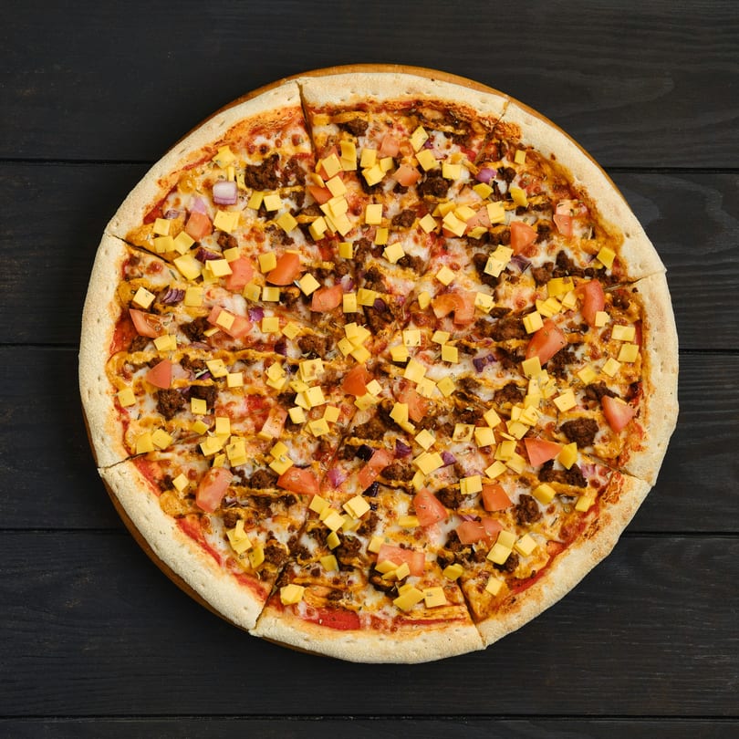 Round pizza with ground meat, diced tomatoes, red onions, and cheese cubes on a dark wooden table.