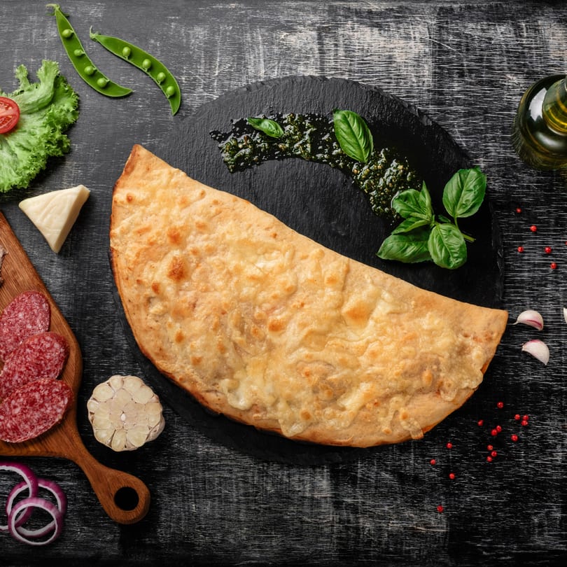 Golden-brown calzone with pesto, basil, salami, cheese, and vegetables on a dark wooden table.