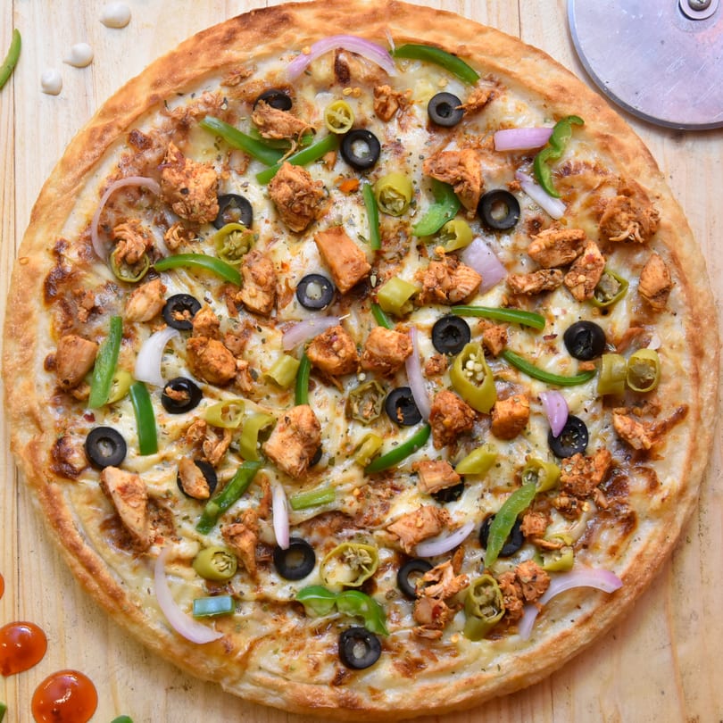 A chicken pizza with olives, green peppers, and onions on a wooden surface with a pizza cutter.
