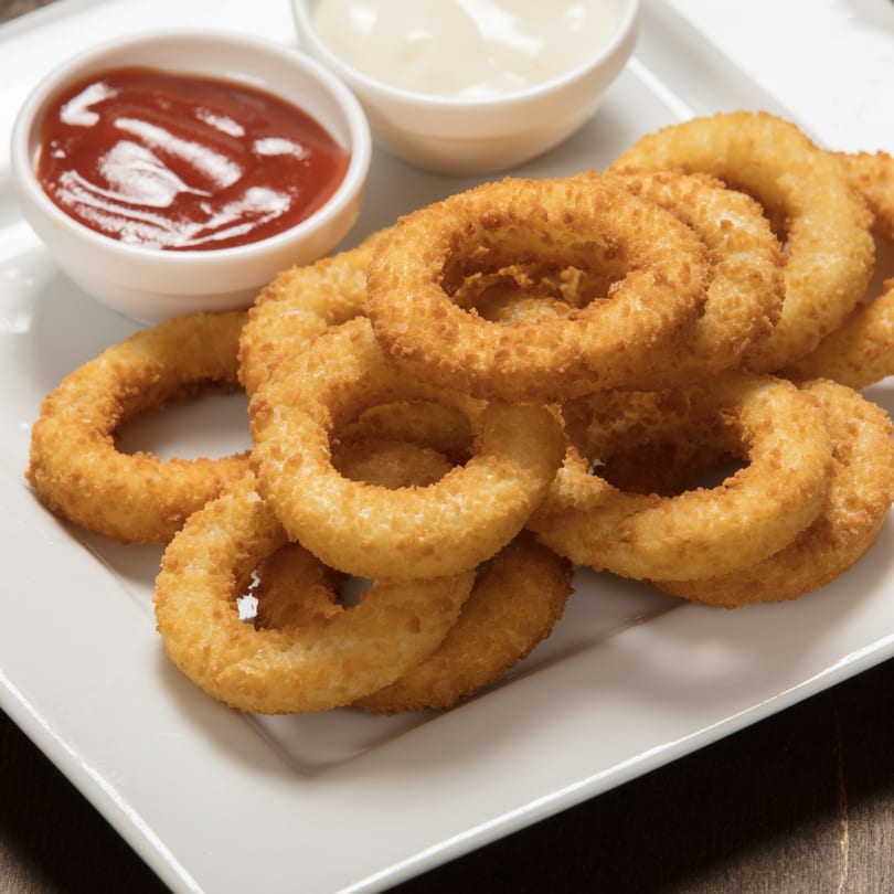Golden-brown onion rings piled on a white plate with small bowls of ketchup and mayonnaise.